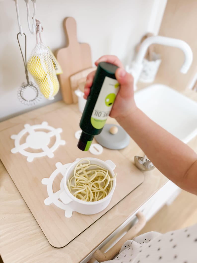 Pu&ograve; includere: Un set da cucina per bambini con un lavello bianco e un piano di lavoro in legno. Una piccola pentola bianca con spaghetti gialli &egrave; sul piano di lavoro. Una bottiglia verde con un'etichetta bianca che dice "110 34" &egrave; tenuta da una mano di bambino. La bottiglia sta venendo versata sugli spaghetti.