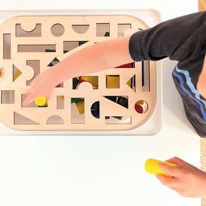 May include: A child's hand reaches into a wooden frame with cutouts of different shapes. The frame is filled with colorful wooden blocks. The child is holding a yellow block in their hand.