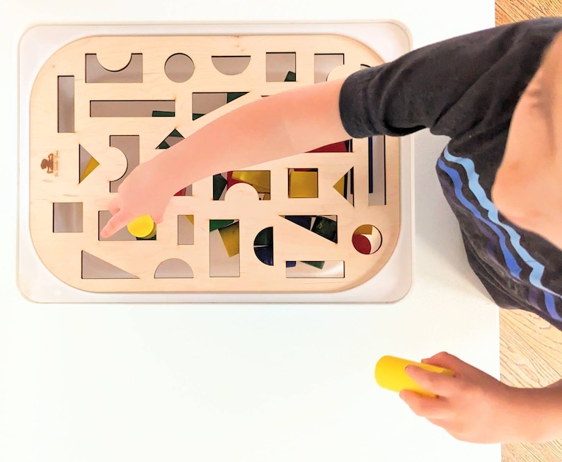 May include: A child's hand reaches into a wooden frame with cutouts of different shapes. The frame is filled with colorful wooden blocks. The child is holding a yellow block in their hand.