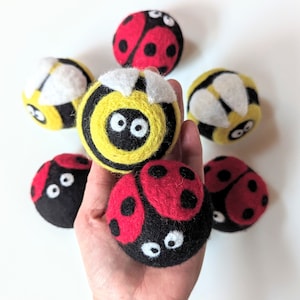May include: A selection of handmade felted wool insect toys, including ladybirds and bumblebees. The ladybirds are red and black with white eyes, while the bumblebees are yellow and black with white accents. These toys are designed to look like insects.
