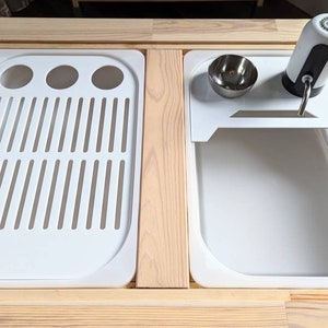 Puede incluir: Una encimera de madera blanca con dos fregaderos empotrados. El fregadero de la izquierda tiene un escurridor de platos blanco con un desag&uuml;e. El fregadero de la derecha tiene un estante blanco con un cuenco peque&ntilde;o y un dispensador de agua.