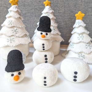 May include: A snowman made of felt with a black top hat and white felt balls. The snowman is sitting on a table with three white felt Christmas trees behind it.
