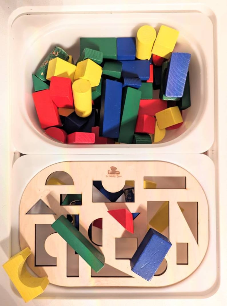 May include: A wooden toy with a cutout shape sorting board and a separate container filled with colorful wooden blocks. The board has a variety of shapes cut out, including circles, squares, triangles, and rectangles. The blocks are red, blue, green, and yellow.