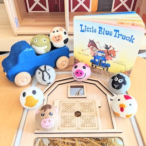 Puede incluir: Un camión de juguete azul con juguetes de animales de fieltro. Los animales incluyen una vaca, un cerdo, una oveja, un pato, una gallina y una rana. El libro "Little Blue Truck" está en el fondo.