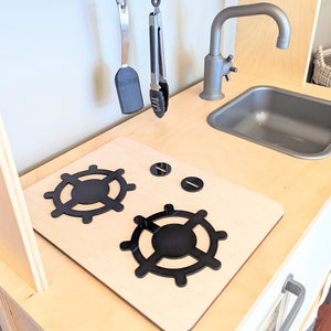 Pu&ograve; includere: Un piano cottura in legno per cucina giocattolo con due manopole nere per i bruciatori e due manopole in metallo nero. Il piano cottura &egrave; in legno chiaro e ha uno sfondo bianco.
