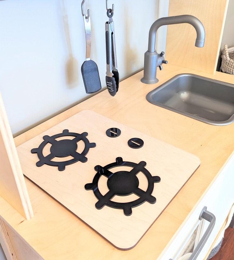Pu&ograve; includere: Un piano cottura in legno per cucina giocattolo con due manopole nere per i bruciatori e due manopole in metallo nero. Il piano cottura &egrave; in legno chiaro e ha uno sfondo bianco.