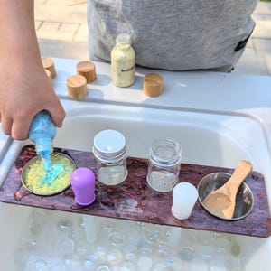 May include: A wooden tray with three glass jars, a purple lid, a white lid, and a small metal bowl with a wooden spoon. The tray is sitting in a sink filled with clear water and iridescent beads.