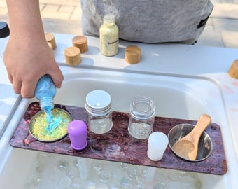May include: A wooden tray with three glass jars, a purple lid, a white lid, and a small metal bowl with a wooden spoon. The tray is sitting in a sink filled with clear water and iridescent beads.