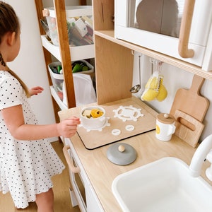 Pu&ograve; includere: Una cucina giocattolo bianca con un piano di lavoro in legno, un lavello bianco, un piano cottura con una pentola e un coperchio e un forno a microonde. Una bambina &egrave; in piedi davanti alla cucina, guardando il piano cottura.