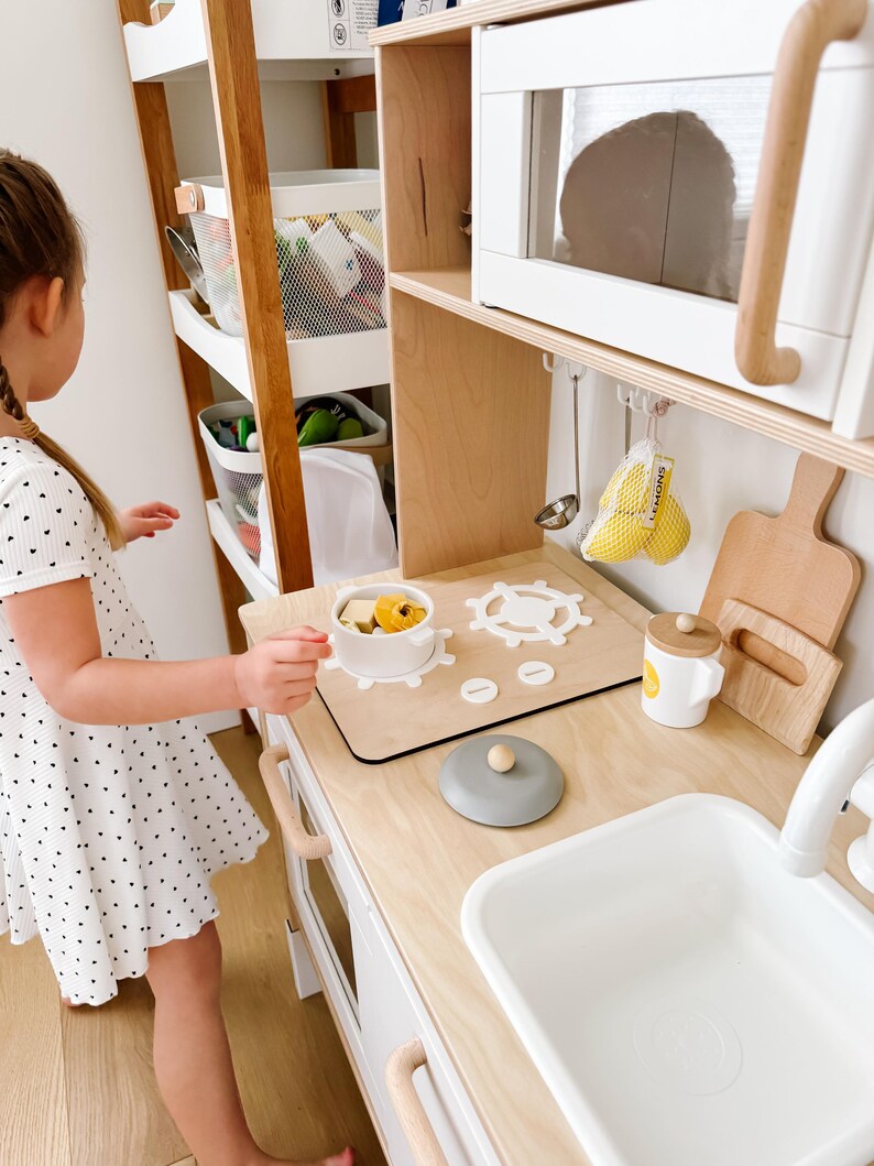 Pu&ograve; includere: Una cucina giocattolo bianca con un piano di lavoro in legno, un lavello bianco, un piano cottura con una pentola e un coperchio e un forno a microonde. Una bambina &egrave; in piedi davanti alla cucina, guardando il piano cottura.