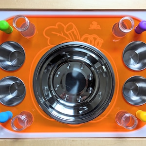 May include: Orange tray with a stainless steel bowl and six smaller bowls. The tray has a white border and a carrot design. There are also six clear glass tubes and six colorful silicone stoppers.
