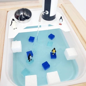 K&ouml;nnte beinhalten: Ein wei&szlig;er sensorischer Wassertisch aus Kunststoff mit hellblauem Wasser, ausgestattet mit einem batteriebetriebenen Wasserspender, mehreren kleinen Pinguinfiguren, wei&szlig;en Schaumstoffbl&ouml;cken und durchsichtigen blauen W&uuml;rfeln im Becken.
