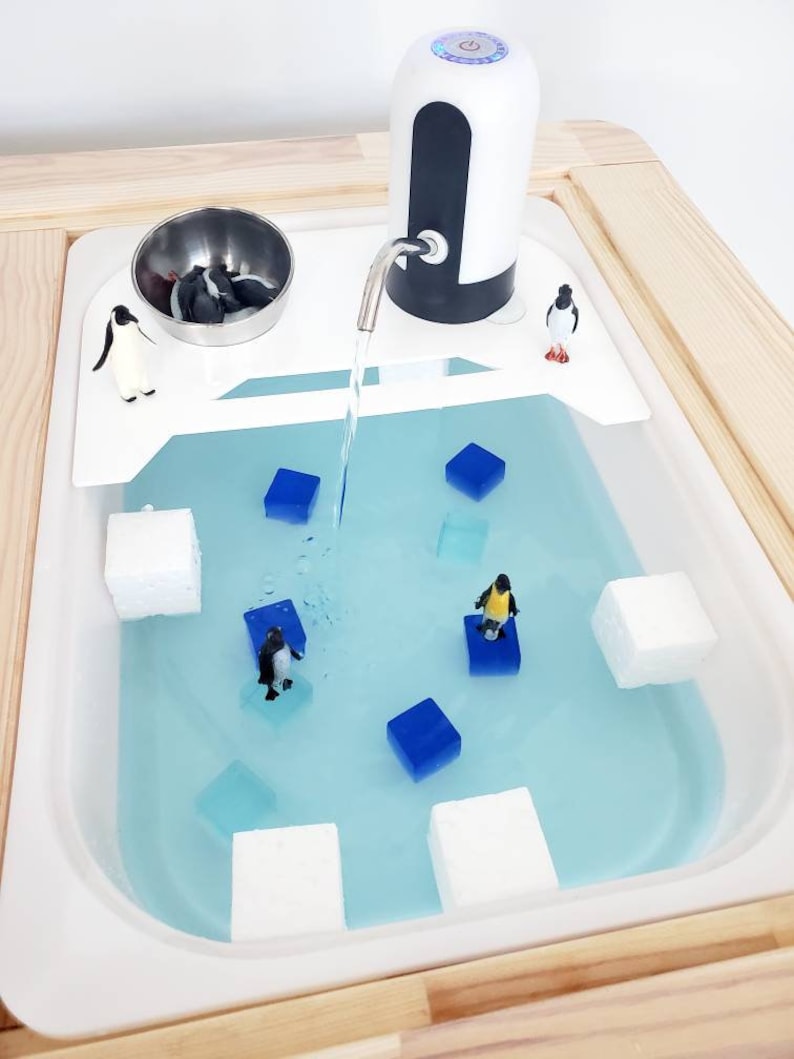 K&ouml;nnte beinhalten: Ein wei&szlig;er sensorischer Wassertisch aus Kunststoff mit hellblauem Wasser, ausgestattet mit einem batteriebetriebenen Wasserspender, mehreren kleinen Pinguinfiguren, wei&szlig;en Schaumstoffbl&ouml;cken und durchsichtigen blauen W&uuml;rfeln im Becken.