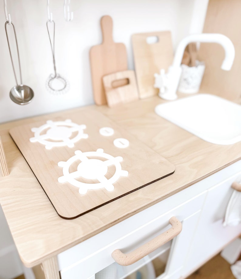 Pu&ograve; includere: Una cucina giocattolo in legno bianco con un lavello bianco e un piano cottura in legno con due manopole bianche per i bruciatori. Il piano cottura ha un piano di lavoro in legno bianco.