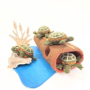 Peut inclure: Quatre tortues en laine feutrée verte avec des motifs de carapace orange et blancs. Trois tortues reposent sur une bûche en feutre marron, tandis qu'une autre est posée sur un rocher clair près d'une base bleue. Une découpe en bois de roseaux.