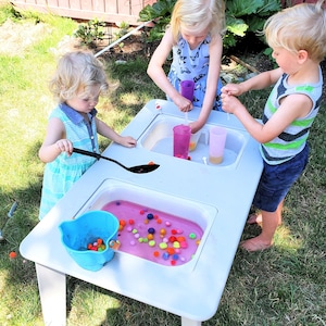 May include: Three children are playing with a white plastic water table that has two basins. One basin has pink water and colourful pom-poms. The other basin has clear water and a blue bucket with pom-poms.