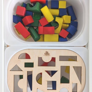 May include: A wooden toy set with a white container filled with colorful wooden blocks and a separate wooden board with cutouts for the blocks. The board has the text "The Wonder Game" printed on it.