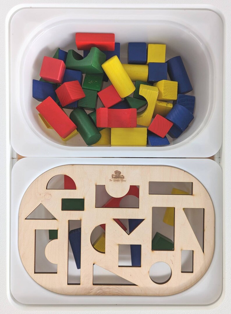 May include: A wooden toy set with a white container filled with colorful wooden blocks and a separate wooden board with cutouts for the blocks. The board has the text "The Wonder Game" printed on it.