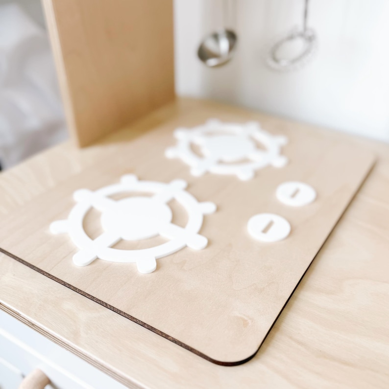 Pu&ograve; includere: Primo piano di un piano cottura da cucina giocattolo in legno con due manopole rotonde bianche. Le manopole hanno un design decorativo che ricorda un timone di nave.