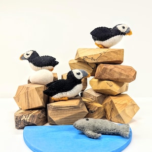 May include: Four felt puffin birds perched on wooden blocks, with a felt grey fish swimming in a blue wooden bowl. The puffin birds are black and white with orange beaks. The wooden blocks are brown and have a natural wood grain.