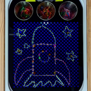 May include: A blue light-up drawing board with a rocket ship design created with colorful pegs. The board has three small containers filled with pegs and a yellow marker labeled "Craft Chalk Writer".