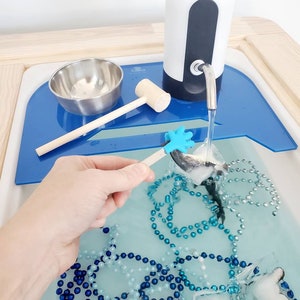 K&ouml;nnte beinhalten: Ein sensorisches Wasserbecken mit einem elektrischen Wasserspender in Wei&szlig; und Dunkelgrau, einer Edelstahlsch&uuml;ssel, einem Holzhammer und einem blauen Kunststoffeinsatz. Das Wasser enth&auml;lt blaue und silberne Perlen sowie Eisw&uuml;rfel mit Spielzeugfiguren.