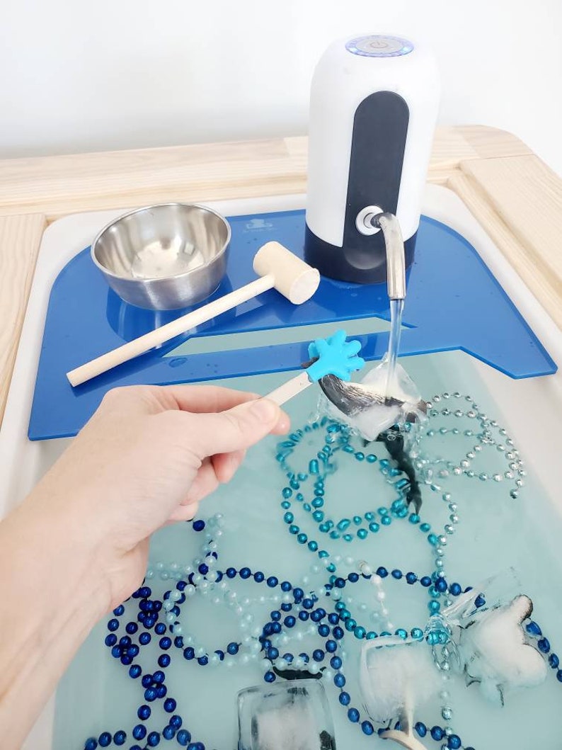 K&ouml;nnte beinhalten: Ein sensorisches Wasserbecken mit einem elektrischen Wasserspender in Wei&szlig; und Dunkelgrau, einer Edelstahlsch&uuml;ssel, einem Holzhammer und einem blauen Kunststoffeinsatz. Das Wasser enth&auml;lt blaue und silberne Perlen sowie Eisw&uuml;rfel mit Spielzeugfiguren.
