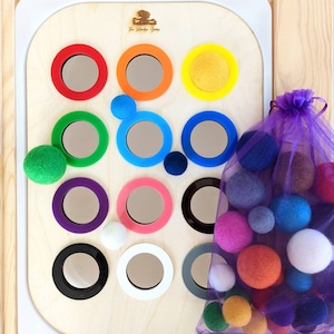 以下が含まれることがあります： 15個のカラフルな円が描かれた木製のボード。各円には鏡が埋め込まれています。ボードの前に、カラフルなフェルトボールでいっぱいの紫色のメッシュバッグがあります。ボードには「The Wonder Years」という文字が印刷されています。