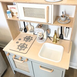Pu&ograve; includere: Un set da cucina giocattolo in legno con un forno a microonde bianco, una macchina per il caff&egrave;, un lavandino e un piano cottura con due fornelli. La cucina ha un armadio grigio chiaro con una porta in vimini intrecciato e un cassetto con una maniglia in legno.