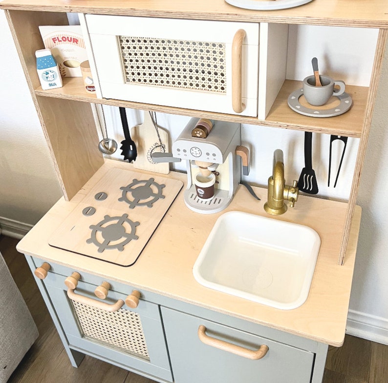 Pu&ograve; includere: Un set da cucina giocattolo in legno con un forno a microonde bianco, una macchina per il caff&egrave;, un lavandino e un piano cottura con due fornelli. La cucina ha un armadio grigio chiaro con una porta in vimini intrecciato e un cassetto con una maniglia in legno.