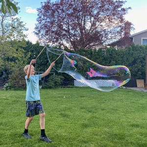 Op de afbeelding: Een jong persoon blaast een grote, regenboogkleurige zeepbel met twee houten stokjes in een grasachtige achtertuin.