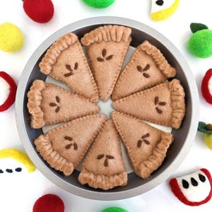 May include: A felt pie set with eight brown felt pie slices in a silver pie pan. The pie slices are surrounded by felt apples and watermelon slices in red, green, and yellow.