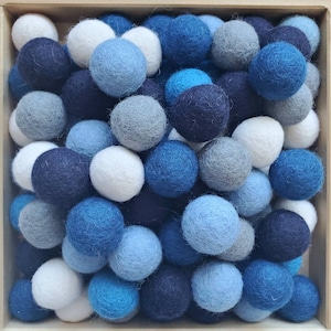 May include: A wooden box overflowing with felt balls in a range of blues, whites, and greys. The balls are tightly packed, offering a tactile and attractive appearance. The colour scheme varies from pale sky blue to dark navy.