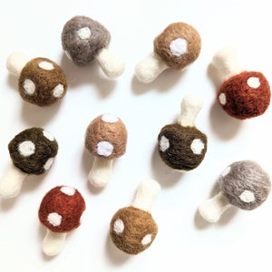 Peut inclure: Dix décorations de champignons en feutre dans différentes nuances de brun, gris et rouge avec des pois blancs. Les champignons ont un pied en feutre blanc.