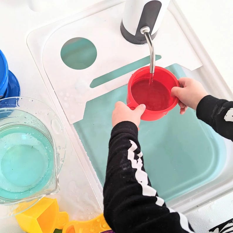 K&ouml;nnte beinhalten: Ein wei&szlig;er Wasser-Spieltisch aus Kunststoff mit einer batteriebetriebenen Wasserpumpe und einem Metallauslauf. Das Set enth&auml;lt einen durchsichtigen Krug mit hellblauem Wasser, einen roten Becher und gelbes Zubeh&ouml;r in einem hellblauen Becken.