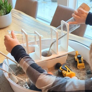 May include: A white wooden structure with clear plastic tubes for pouring sand into a white plastic sandbox. The sandbox is filled with sand and includes toy construction vehicles.
