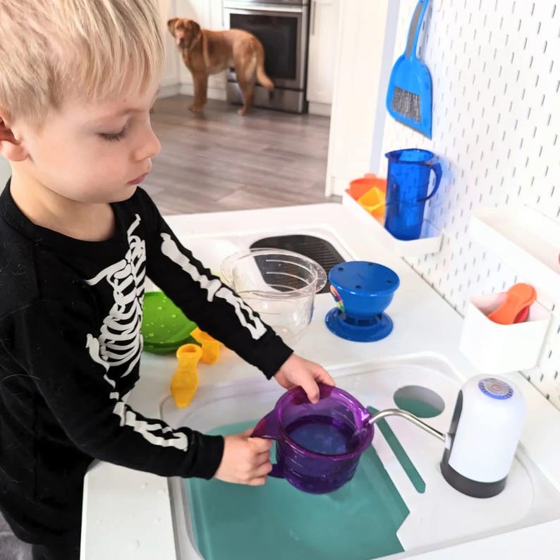 K&ouml;nnte beinhalten: Eine wei&szlig;e Spielk&uuml;chen-Sp&uuml;le aus Kunststoff mit einem t&uuml;rkisfarbenen Becken, einer batteriebetriebenen Wasserpumpe mit Metallauslauf und verschiedenen bunten Kunststoff-Accessoires wie einem lila Messbecher, einem blauen Krug und einer gr&uuml;nen Sch&uuml;ssel.