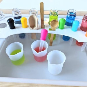 May include: A white plastic sensory play tray insert featuring a curved rack with multiple circular cutouts holding clear glass vials filled with colored liquids, colorful silicone droppers, a wooden scoop, and a wooden spoon, with three translucent silicone beakers containing colored liquids placed below.