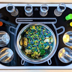 Puede incluir: Una bandeja negra con una olla plateada llena de confeti verde, azul y amarillo y monedas de oro. Hay seis frascos de vidrio transparente y seis tazas de plata alrededor de la olla.