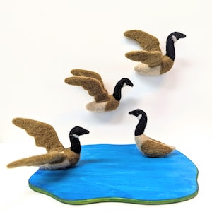 May include: Three felt geese, two in flight and one landing on a blue painted wooden pond. The geese are brown with black heads and white bellies.