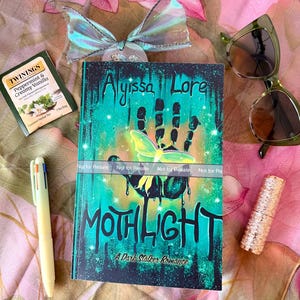 May include: A book titled "Mothlight: A Dark Stalker Romance" with a teal and black cover, a pen, sunglasses, a tea bag, and a decorative bow. The book cover features a handprint and a moth illustration. The background is a floral pattern.