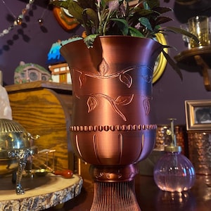 May include: A copper-toned decorative vase featuring a fluted base and a detailed leaf and vine pattern. The vase contains a small plant with green and purple leaves, adding a natural element to the ornate design.