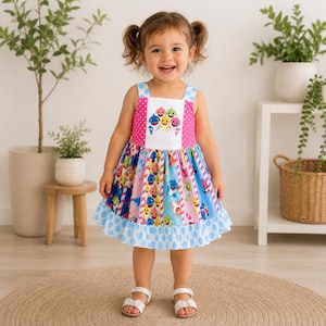 Può includere: Un vestito colorato per bambini piccoli a tema 'Baby Shark'. L'abito ha un corpetto rosa con pois bianchi, uno scollo quadrato e una gonna con vari personaggi di squali. L'orlo è rifinito con pois blu. La bambina indossa sandali bianchi.