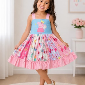 May include: A young girl wearing a pink and blue dress with a Peppa Pig graphic. The dress has a pink ruffle hem and straps with pink polka dots. She is also wearing white sandals and a pink bow in her hair.