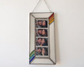 May include: A rectangular stained glass photo frame with a white border and a metal chain for hanging. The frame features decorative corner accents in red, orange, and yellow at the top right, and green and blue at the bottom left, holding a photo strip.