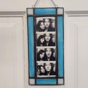 May include: A rectangular stained glass photo frame with a light blue border and a chain for hanging. The frame holds four black and white photo booth style images of a couple. The frame is hanging on a white door.