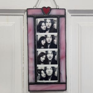 May include: A stained glass photo frame with a pink and purple border. The frame holds four black and white photo booth style images of a couple. A red heart is at the top, and the frame hangs from a chain.