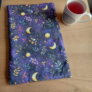 Può includere: Una custodia in tessuto rettangolare con un motivo celeste blu e viola scuro caratterizzato da lune crescenti, lune piene, stelle gialle e vari rametti botanici. La custodia presenta un piccolo bottone a pressione viola sul bordo superiore.