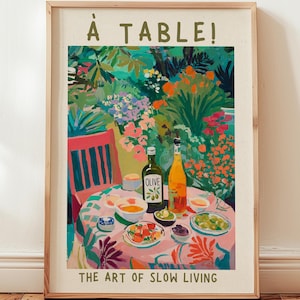 Może przedstawiać: Oprawiony wydruk artystyczny z kolorową ilustracją stołu nakrytego do posiłku w ogrodzie. Tekst głosi À TABLE! oraz THE ART OF SLOW LIVING. Scena przedstawia butelkę oliwy z oliwek, butelkę wina oraz miski z jedzeniem na obrusie w kwiatowe wzory.