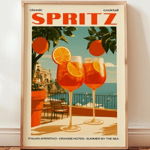 Könnte beinhalten: Ein gerahmter Kunstdruck mit einer klassischen Spritz-Cocktail-Werbung. Das Bild zeigt zwei Gläser des orangefarbenen Getränks, garniert mit Orangenscheiben, vor einer Küstenkulisse mit Orangen und dem Text "Italian Aperitivo - Orange Notes - Summer by the Sea."